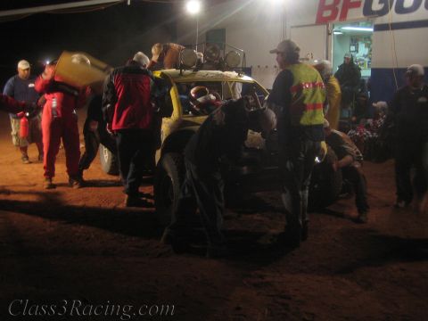 1990 Ford Bronco - Crew refuel a yellow off-road race buggy with jugs at a busy night pit lit by overhead lamps and a BFGoodrich banner.