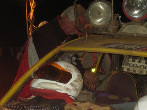 1990 Ford Bronco - gray-haired crew member leans over the front of a yellow off-road race car at night as a white helmet rests on the body.