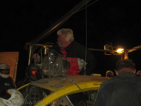 1990 Ford Bronco - gray-haired man in a red and black jacket reaches into the rear of a yellow off-road race car at night near helmets and a light.