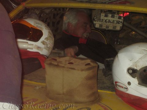 1990 Ford Bronco - smiling gray-haired man reclines inside a yellow off-road race car next to a tan fuel cell and white racing helmets.
