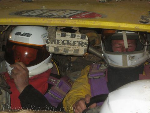 1990 Ford Bronco - Two drivers in white racing helmets sit inside a mud-caked yellow off-road race car, a Checkers Off-Road Racing Team sticker visible behind…