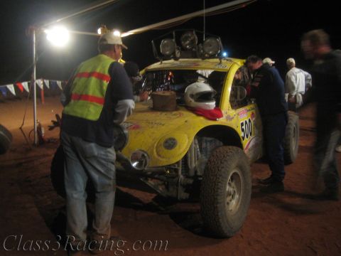 1990 Ford Bronco - man in a reflective vest stands beside a yellow off-road race car numbered 500 with roof-mounted lights, parked on red dirt at a night pit…