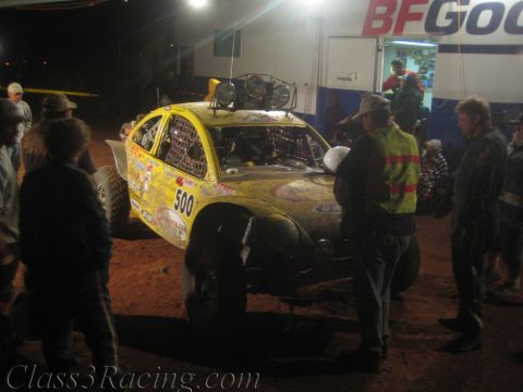 1990 Ford Bronco - Crew members in vests gather around yellow off-road race car #500 in a dirt pit area at night, with a BFGoodrich trailer behind.