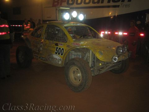 1990 Ford Bronco - Yellow off-road race car number 500 with roof lights sits on red dirt in front of a BFGoodrich Tires banner during a night event.
