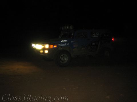 1990 Ford Bronco - dark blue off-road race truck numbered 760 drives across the desert at night with its headlights and roof light bar illuminated.