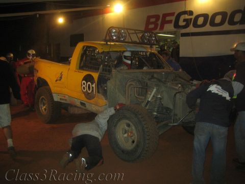 1990 Ford Bronco - yellow off-road race truck numbered 801 sits with its rear suspension exposed as crew members work on it under a BFGoodrich banner at night.