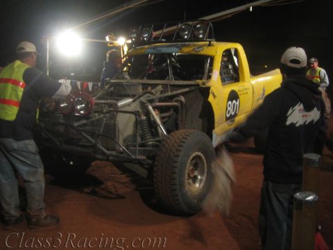 1990 Ford Bronco - Crew members push yellow off-road race truck number 801 with its rear chassis and shocks exposed across red dirt at a night pit stop.