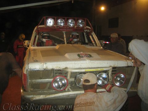 1990 Ford Bronco - weathered white off-road race truck with four round roof lights is surrounded by crew members during a nighttime pit stop.