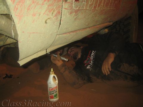1990 Ford Bronco - man lies on the dirt beneath a scraped race car working with a tool, a Castrol gear oil bottle on the ground beside him.