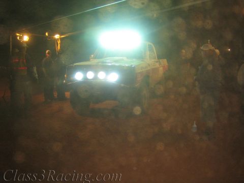 1990 Ford Bronco - off-road race truck glows under a bright roof light bar amid heavy dust, with crew silhouettes standing nearby at night.