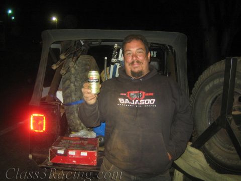 1990 Ford Bronco - smiling man wearing a Baja 500 hoodie holds up a beer can at the open rear of a vehicle with a spare tire, at night.
