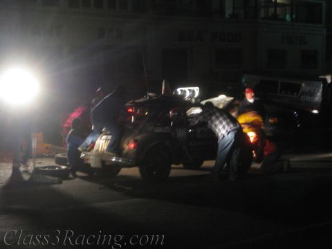 1990 Ford Bronco - group of people work on a race car parked on pavement in front of a motel and bar at night, with a bright floodlight to the side.