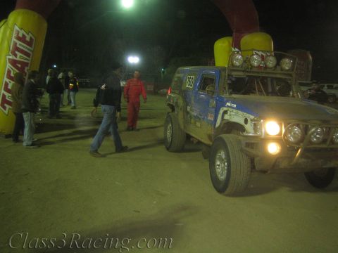 1990 Ford Bronco - blue off-road race truck numbered 760 with headlights on drives past a yellow-and-red Tecate inflatable arch as spectators watch at night.