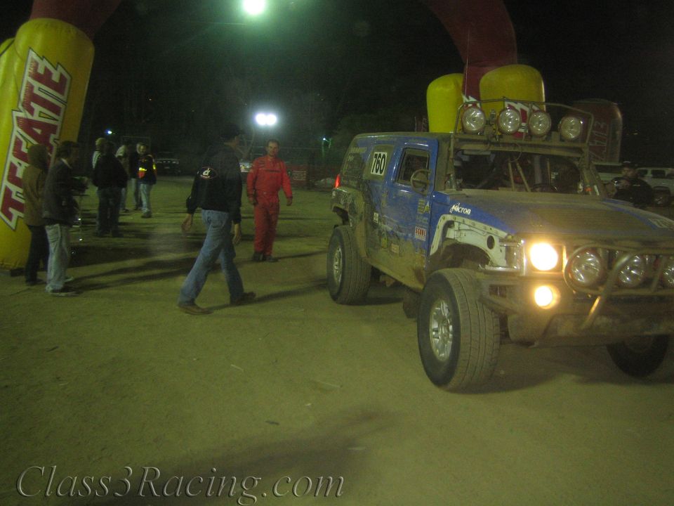 Blue off-road race truck 760 passing under a Tecate inflatable arch at night