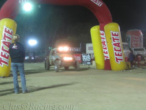 1990 Ford Bronco - spectator watches an off-road vehicle with bright lights approach a large red Tecate inflatable finish arch at a night event.