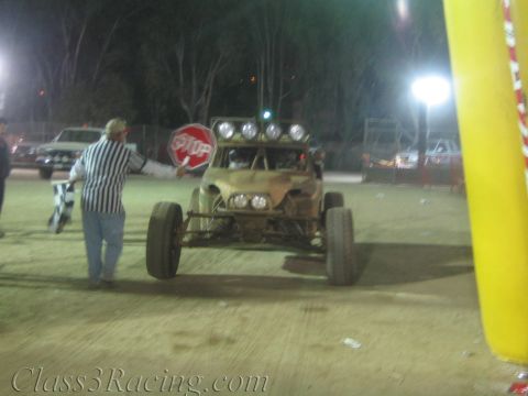 1990 Ford Bronco - official in a striped shirt holds a 5-Up stop sign beside a tan open-wheel off-road buggy on a dirt staging area at night.