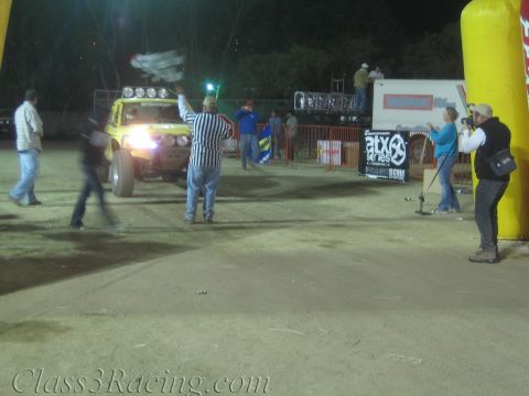 1990 Ford Bronco - striped-shirt official raises a flag to launch a yellow off-road race vehicle while spectators and photographers watch at the dirt start li…