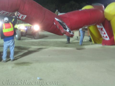 1990 Ford Bronco - red Tecate inflatable arch has partially collapsed over the dirt course as a crew member in a reflective vest stands nearby at night.