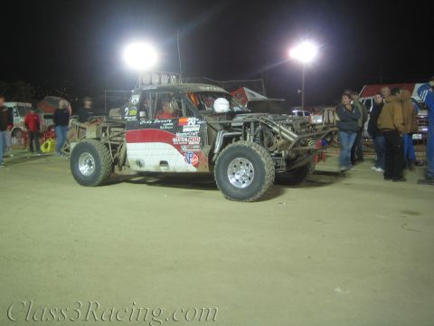 1990 Ford Bronco - white-and-red tube-chassis off-road race car numbered 2 sits under stadium lights on a dirt pit area as spectators stand around it at night.