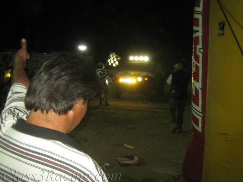 1990 Ford Bronco - Over the shoulder of a striped-shirt spectator, an off-road truck with a bright light bar approaches a flagger at the dirt finish at night.