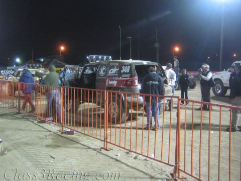 1990 Ford Bronco - dark Toyota off-road race truck numbered 348 with Donahoe Racing markings sits behind orange crowd fencing among other vehicles at night.