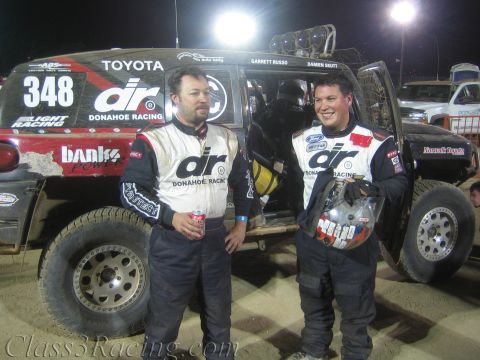 1990 Ford Bronco - Two men in Donahoe Racing suits stand smiling beside their Toyota off-road race truck number 348 under floodlights at night.