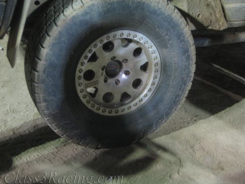 1990 Ford Bronco - mud-coated off-road tire mounted on a white beadlock alloy wheel, photographed beneath a truck.