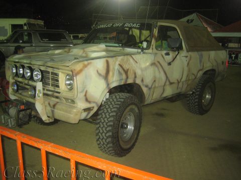 1990 Ford Bronco - lifted SUV in tan-and-brown camouflage paint with a front light pod sits behind an orange barrier at night.
