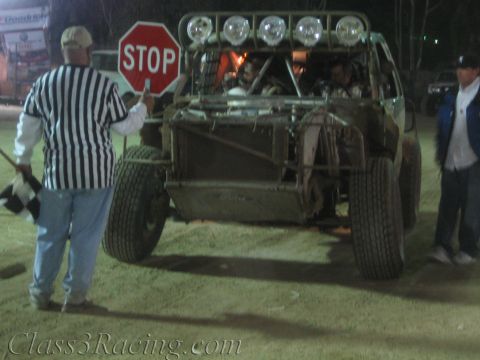 1990 Ford Bronco - striped-shirt official raises a STOP sign before a roll-caged off-road race vehicle with five roof lights at night.