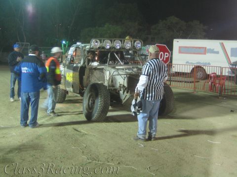 1990 Ford Bronco - roll-caged off-road race vehicle waits in a dirt staging area as officials and spectators stand nearby at night.