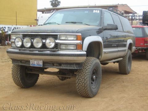1990 Ford Bronco - black lifted Chevrolet Suburban with a four-light front bar and California plate sits on a dirt lot.