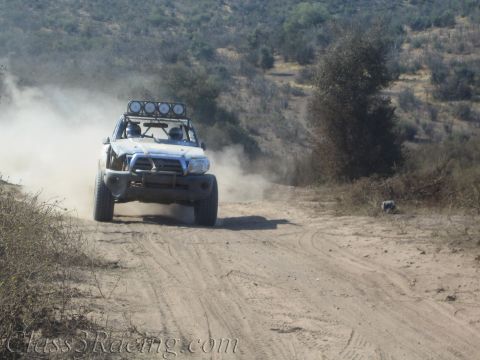1990 Ford Bronco - white off-road prerunner truck with a roof light bar races down a dirt trail throwing up a dust cloud.