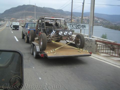 1990 Ford Bronco - roll-caged off-road buggy strapped to a flatbed trailer is towed across a bridge above a reservoir.