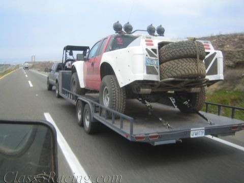 1990 Ford Bronco - red-and-white off-road race truck carrying spare tires is towed on a flatbed trailer along a highway.