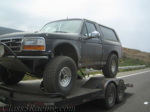 1990 Ford Bronco - black Ford Bronco with a rear hardtop sits lifted on a flatbed trailer beside a coastal highway.