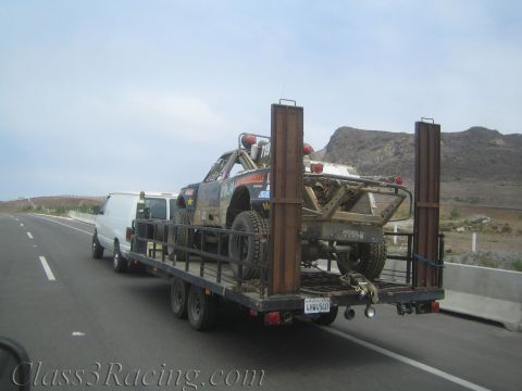 1990 Ford Bronco - roll-caged off-road race truck loaded on a flatbed trailer is towed by a white van along a desert highway.