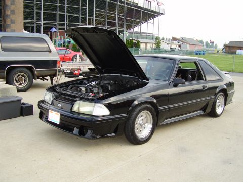 1988 Ford Mustang - A black 1987 Ford Mustang is parked outside with its hood open, next to a two-tone SUV.