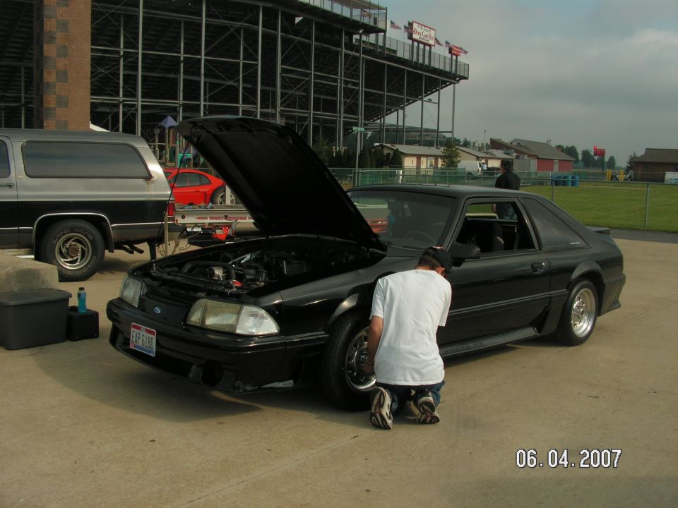 Black coupe with hood open, person kneeling by front wheel. Silver SUV parked to the left. Grandstands in background