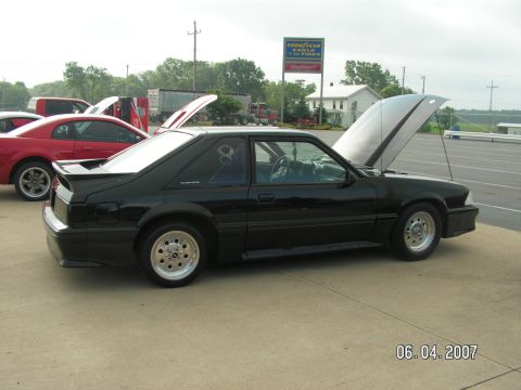 1988 Ford Mustang - A black coupe sits with its hood open, parked next to a red car on a paved lot.