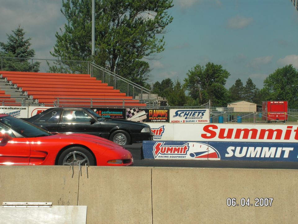 Black Mustang and red Corvette at drag strip, grandstands in background