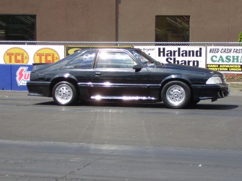 1988 Ford Mustang - A black coupe with racing wheels is seen from the side on a track.