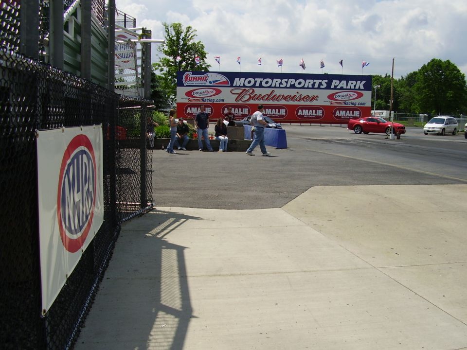 Drag strip scene with a red car and white van, spectators, and a large motorsports park banner