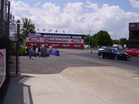 1988 Ford Mustang - A dark blue coupe drives down the track at Motorsports Park, with spectators visible near the grandstands.