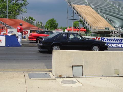 1988 Ford Mustang - A black coupe and a red convertible prepare for a drag race at a track with grandstands.