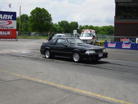 1988 Ford Mustang - A black 1988 Ford Mustang is seen on the drag strip, with an ambulance and other vehicles in the background.