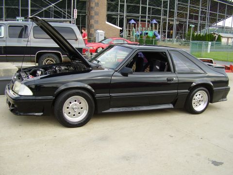 1988 Ford Mustang - A black coupe with its hood open sits on pavement, with a grey van and a red car in the background.
