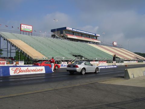 1988 Ford Mustang - Two cars prepare for a drag race at a motorsports park with large spectator grandstands.