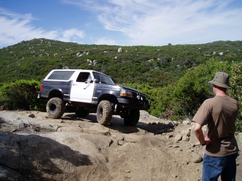 1994 Ford Bronco - A two-tone Ford Bronco navigates a rocky off-road path under a partly cloudy sky.