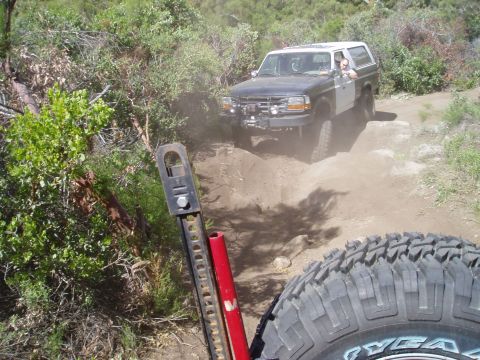 1994 Ford Bronco - Two SUVs navigate a dusty, off-road trail surrounded by dense vegetation and rocky terrain.