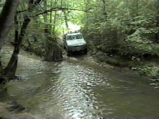 1980 Ford Bronco - SUV off-roading through a stream in a dense forest setting. Supermotors.net branding is present.